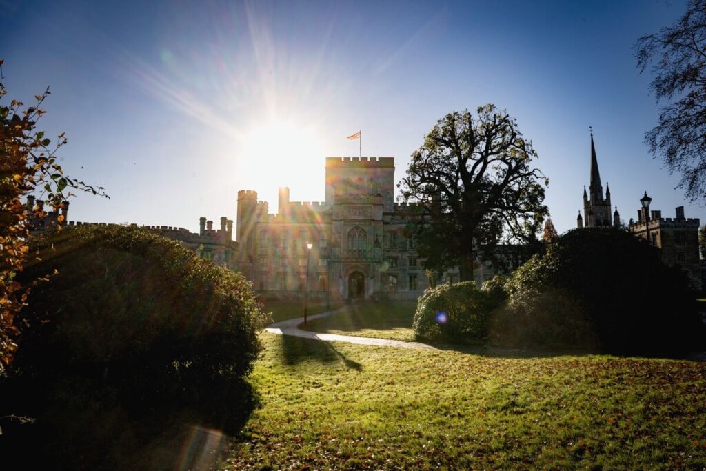 The sun shines brightly behind a historic castle with tall towers and a flag, casting long shadows over the green lawn and bushes. A church steeple rises in the background under a clear blue sky.