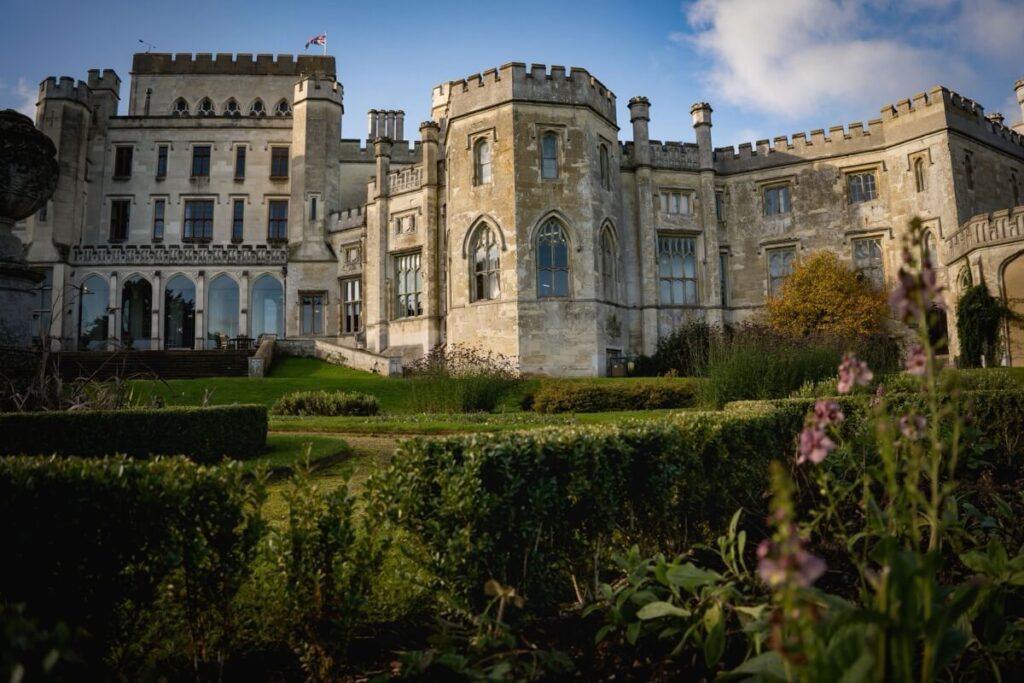 A large, historic stone castle with turrets and arched windows stands behind well-manicured gardens, with flowers and hedges in the foreground under a partly cloudy sky.