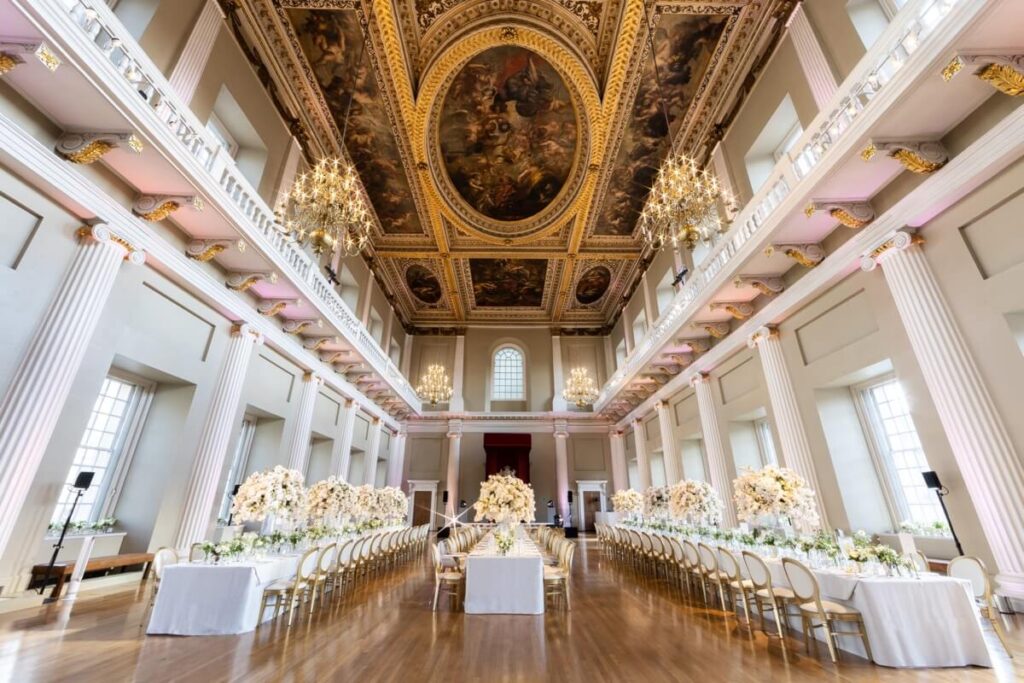 A grand hall set for a formal event, featuring long banquet tables with elegant white floral centerpieces, high ceilings with ornate paintings, large windows, chandeliers, and classical columns.