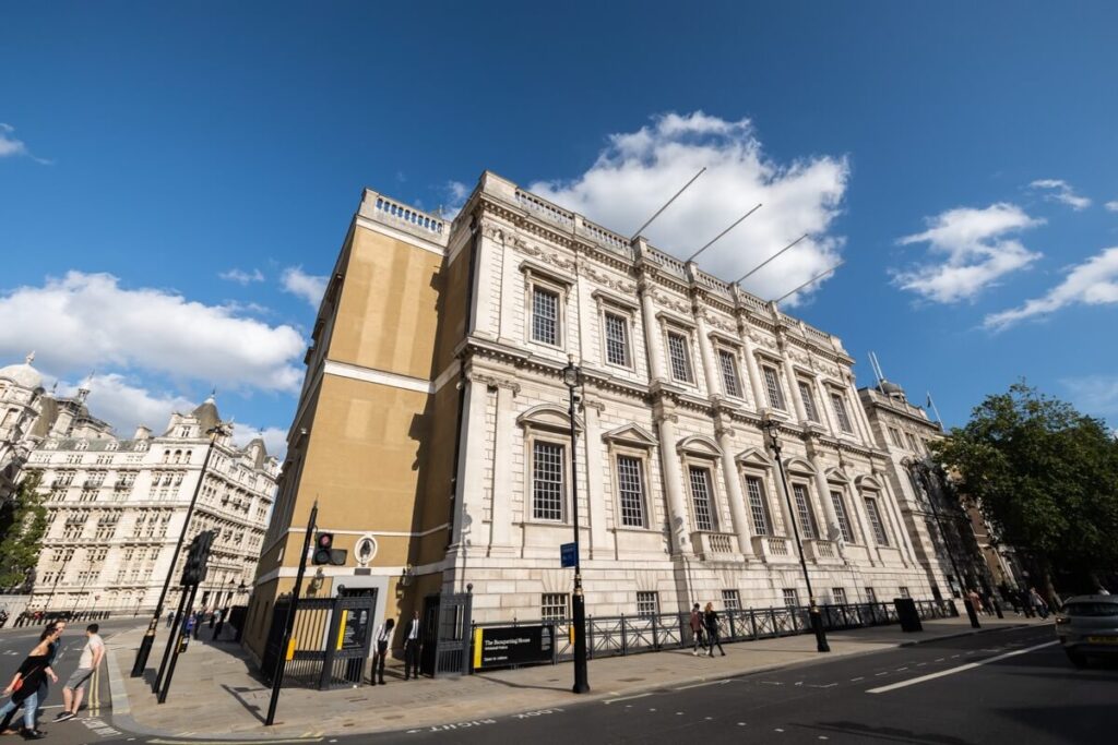 A large neoclassical building with tall windows and ornate stonework stands on a city corner under a blue sky with scattered clouds; several people walk along the sidewalk.