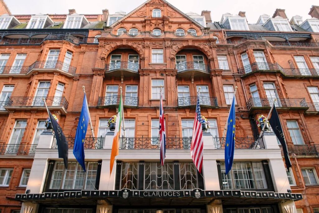 The facade of Claridge’s hotel, a historic red-brick building with balconies and arched windows, featuring several international flags displayed above the entrance.
