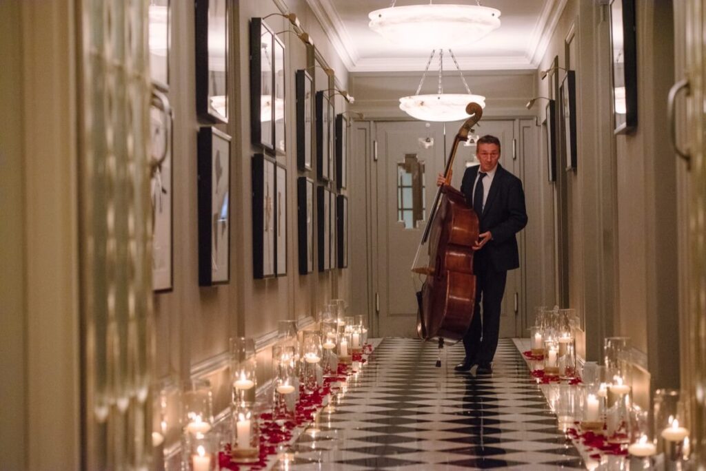 A man in a suit stands in a hallway holding a double bass. The hallway is lined with lit candles, flower petals, and framed pictures on the walls, creating a romantic and elegant atmosphere.