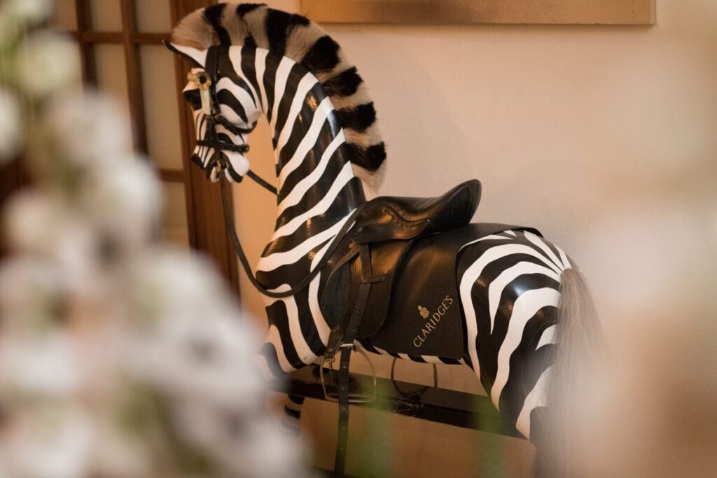 A black and white striped rocking horse designed to look like a zebra, with a black saddle labeled "CLARIDGE'S," positioned indoors against a neutral wall.