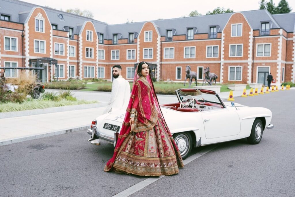 A man in a white outfit and a woman in a red and gold traditional dress stand beside a white vintage convertible car, parked in front of a large brick building with tall windows and neatly trimmed landscaping.