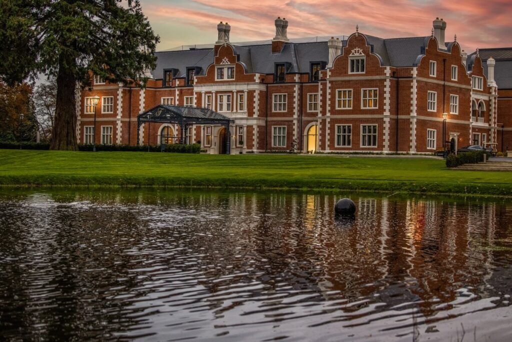A large, elegant red-brick mansion with white trim and chimneys stands behind a pond, reflecting its image. The sky is streaked with warm sunset colors, and green lawns surround the building.