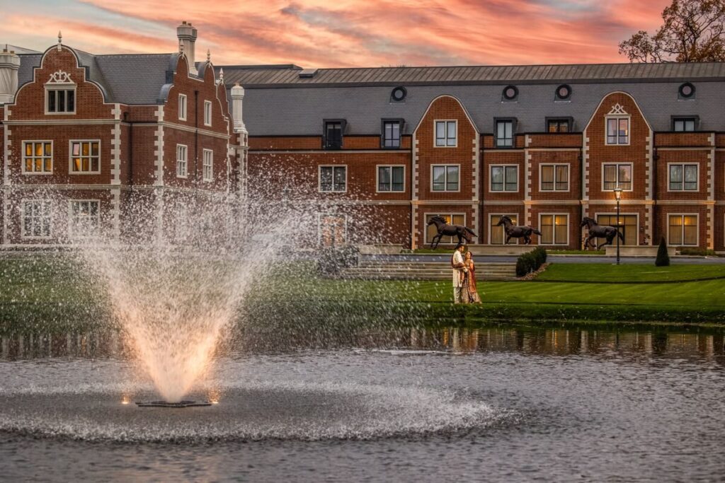 A large water fountain sprays in a pond before a grand brick building at sunset. Two people stand together on the grass near the water, with horse statues visible in the background.
