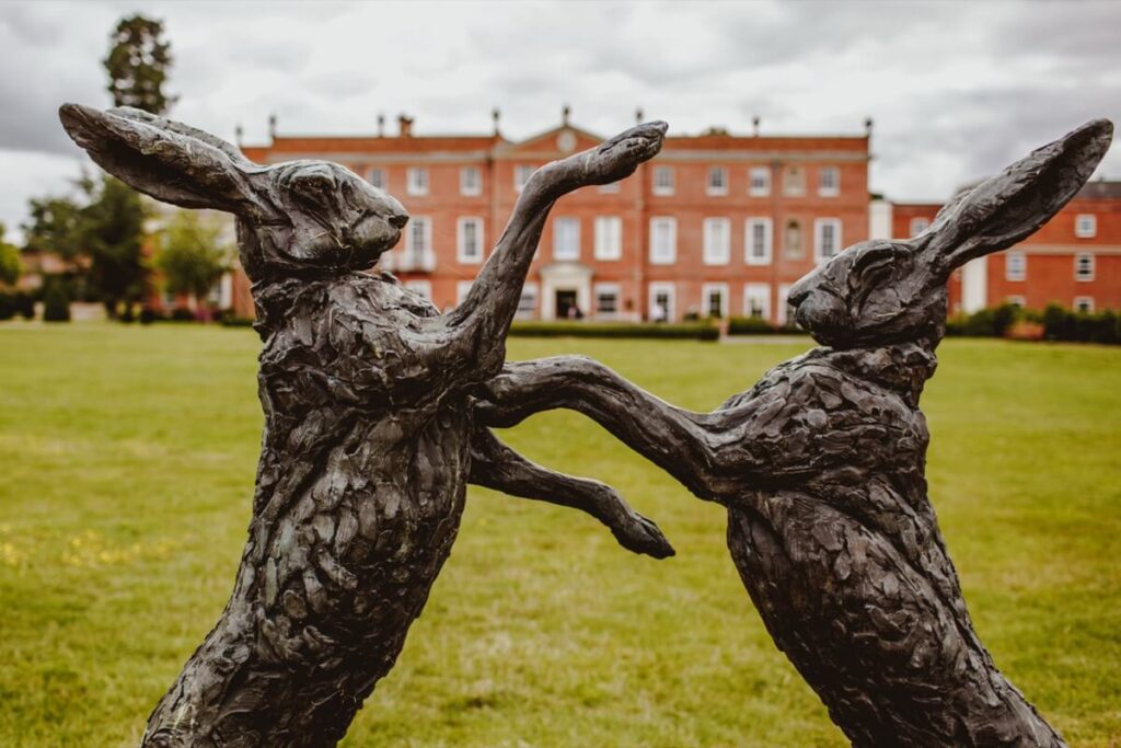 Bronze statue of two hares boxing in a grassy area, with a large red brick building and cloudy sky in the background.