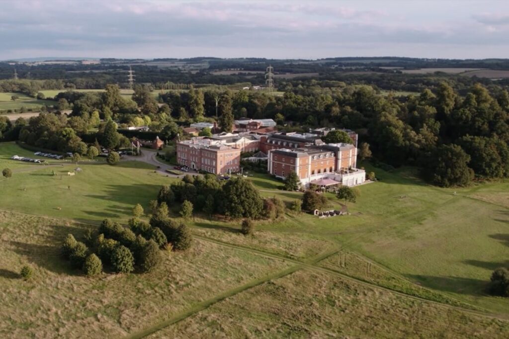 A large, historic brick building surrounded by expansive green fields and trees, viewed from above on a partly cloudy day, with rolling countryside stretching into the distance.