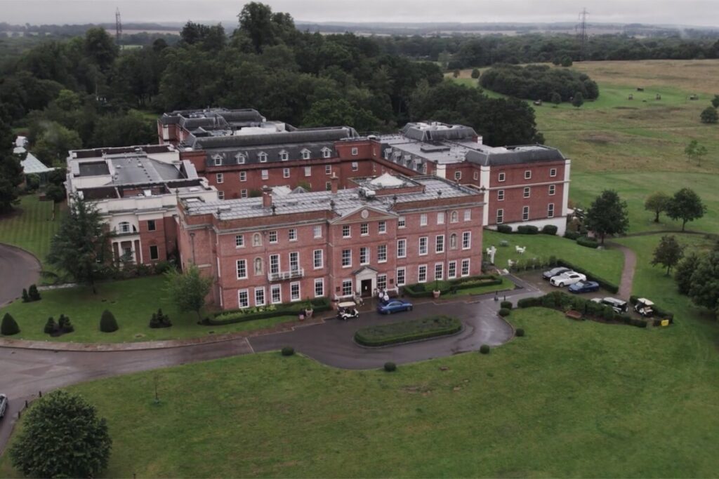 Aerial view of a large, historic red-brick building with white trim, surrounded by lawns, trees, and parked cars, set in a rural landscape with open fields and distant trees.