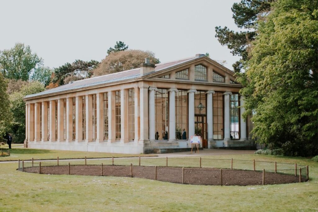 A large glasshouse with tall columns and many windows stands in a park surrounded by green trees and grass, with a small fenced garden bed in the foreground.