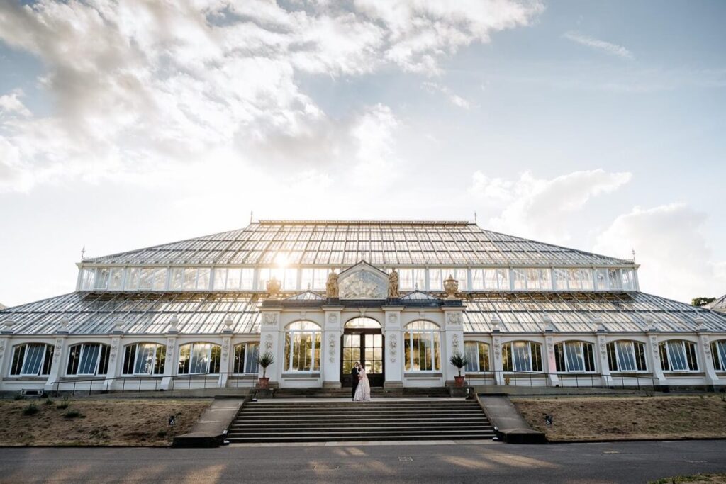 A large glass conservatory is shown at sunset, with the sun shining through the windows. A couple stands at the entrance on the front steps, surrounded by potted plants and greenery. The sky is partly cloudy.