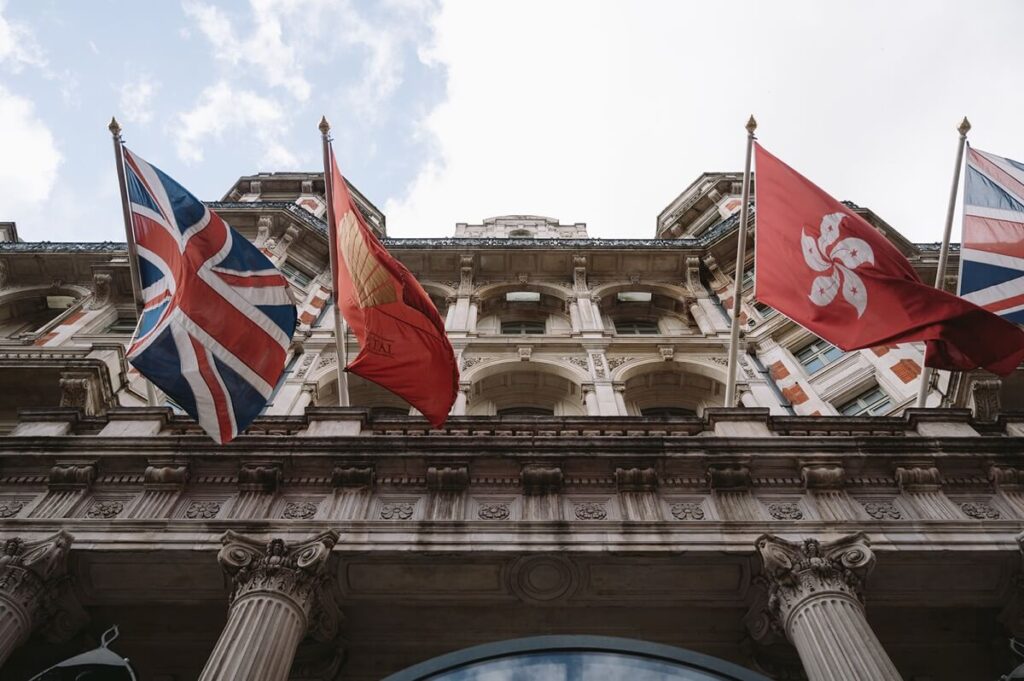 A low-angle view of a historic building with multiple columns, displaying the United Kingdom, Hong Kong, and other flags on flagpoles against a partly cloudy sky.