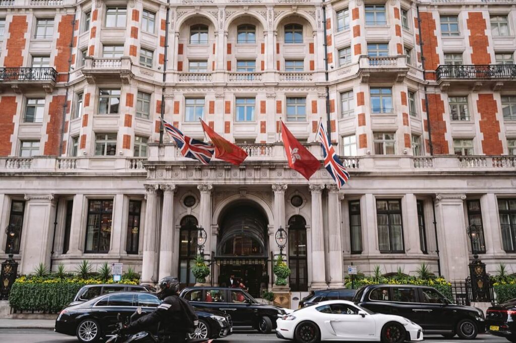 A grand hotel exterior with British and red flags above the entrance. Luxury cars, including a white sports car and black sedans, are parked in front, while a person on a motorcycle rides past. The building has ornate architecture.
