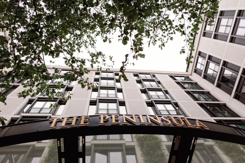 View from below of The Peninsula hotel entrance with gold lettering, framed by modern building façades and leafy branches above.