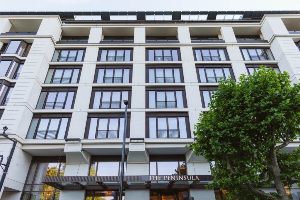 A multi-story, modern hotel building with large windows and balconies. The sign above the entrance reads “The Peninsula.” A leafy tree is in the foreground on the right side of the image.