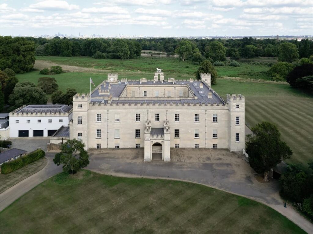 A large, rectangular, light-colored historic building with battlements sits surrounded by expansive green lawns and trees under a partly cloudy sky.