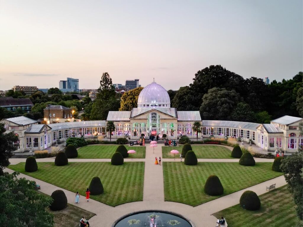 Aerial view of a large glass-domed conservatory surrounded by symmetrical gardens, manicured lawns, garden paths, and trimmed bushes, with people walking and mingling outside at sunset.