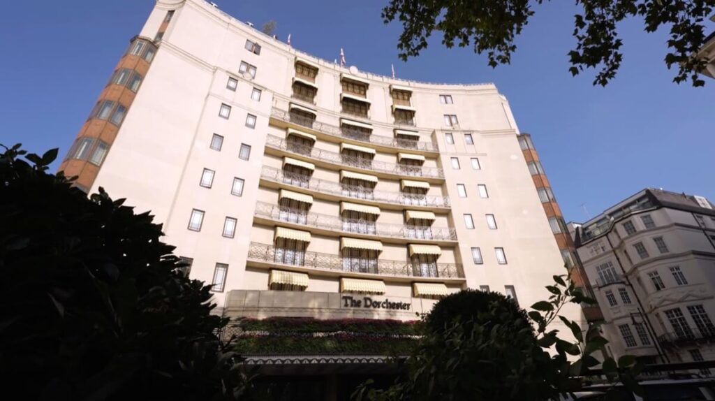 A view of The Dorchester hotel’s exterior, showing its cream-colored facade, decorative balconies, and entrance canopy, framed by trees and greenery under a clear blue sky.