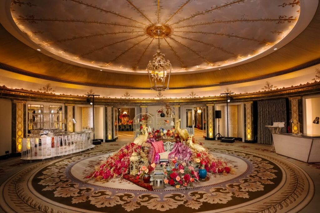 A luxurious, circular room with an ornate ceiling and chandelier. The center features an elaborate floral arrangement with red, pink, and peach flowers, gold chairs, and rich fabrics, surrounded by elegant decor and soft lighting.
