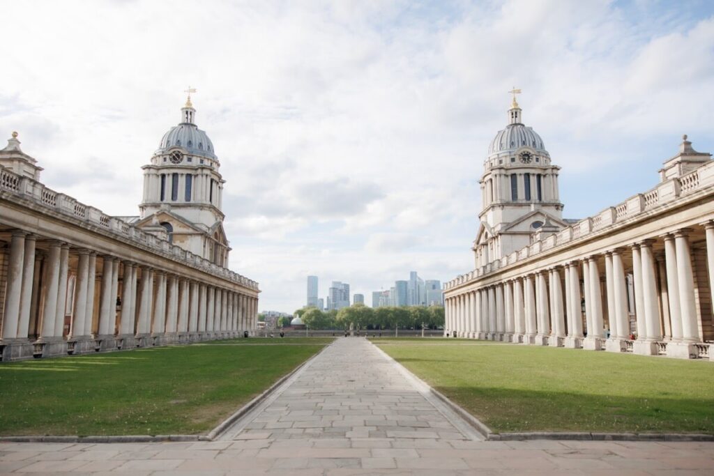 A symmetrical view of two grand historic buildings with domed towers and colonnades, separated by a wide stone path and lawn, with a modern city skyline visible in the distance under a partly cloudy sky.