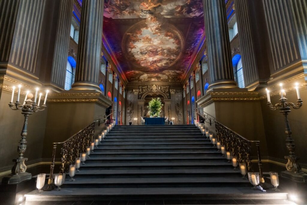Grand staircase in an ornate hall with tall columns, candlelit steps, and elaborate ceiling artwork. At the top, a table with a green floral arrangement stands before a decorated wall.