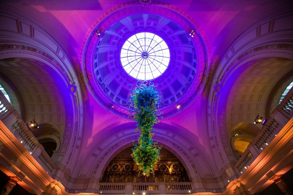 A view looking up at a domed glass ceiling with purple lighting, ornate arches, and a large green glass sculpture hanging from the center.
