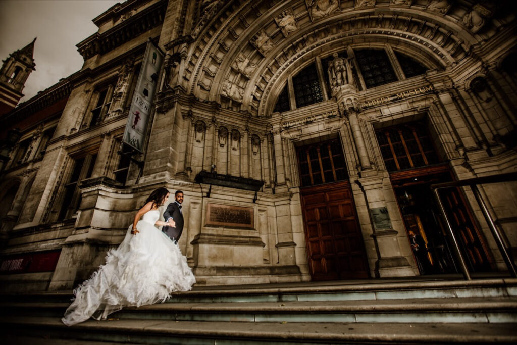 A bride and groom in wedding attire stand on the steps of an ornate, historic building with large wooden doors and intricate stonework. The bride's dress cascades down the stairs as they look at each other.