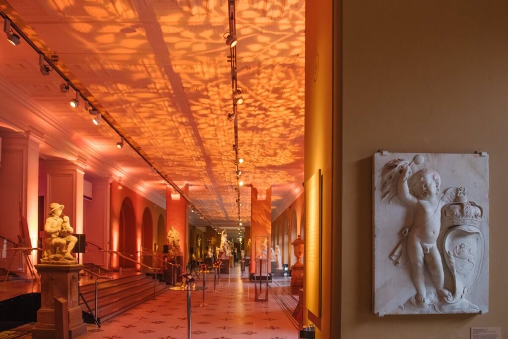 A museum hallway with warm orange lighting, decorative ceiling patterns, sculptures along the walls, and a white relief of a cherub on the right wall in the foreground.